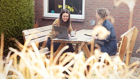 Zwei Frauen sitzen an einem Gartentisch und lachen, während sie einen Laptop betrachten.