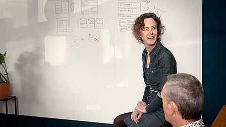 Eine Frau sitz vor einem Whiteboard auf das ein Wireframe gezeichnet wurde. Vor ihr sitzt ein Mann. 